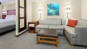 A hotel room with a bed, a gray sofa, a brown ottoman, and a painting on the wall.