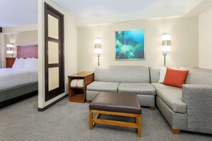 A hotel room with a bed, a gray sofa, a brown ottoman, and a painting on the wall.