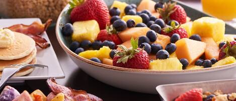 A breakfast spread with various dishes including scrambled eggs, bacon, pancakes, and a fruit bowl with strawberries, blueberries, and pineapple.