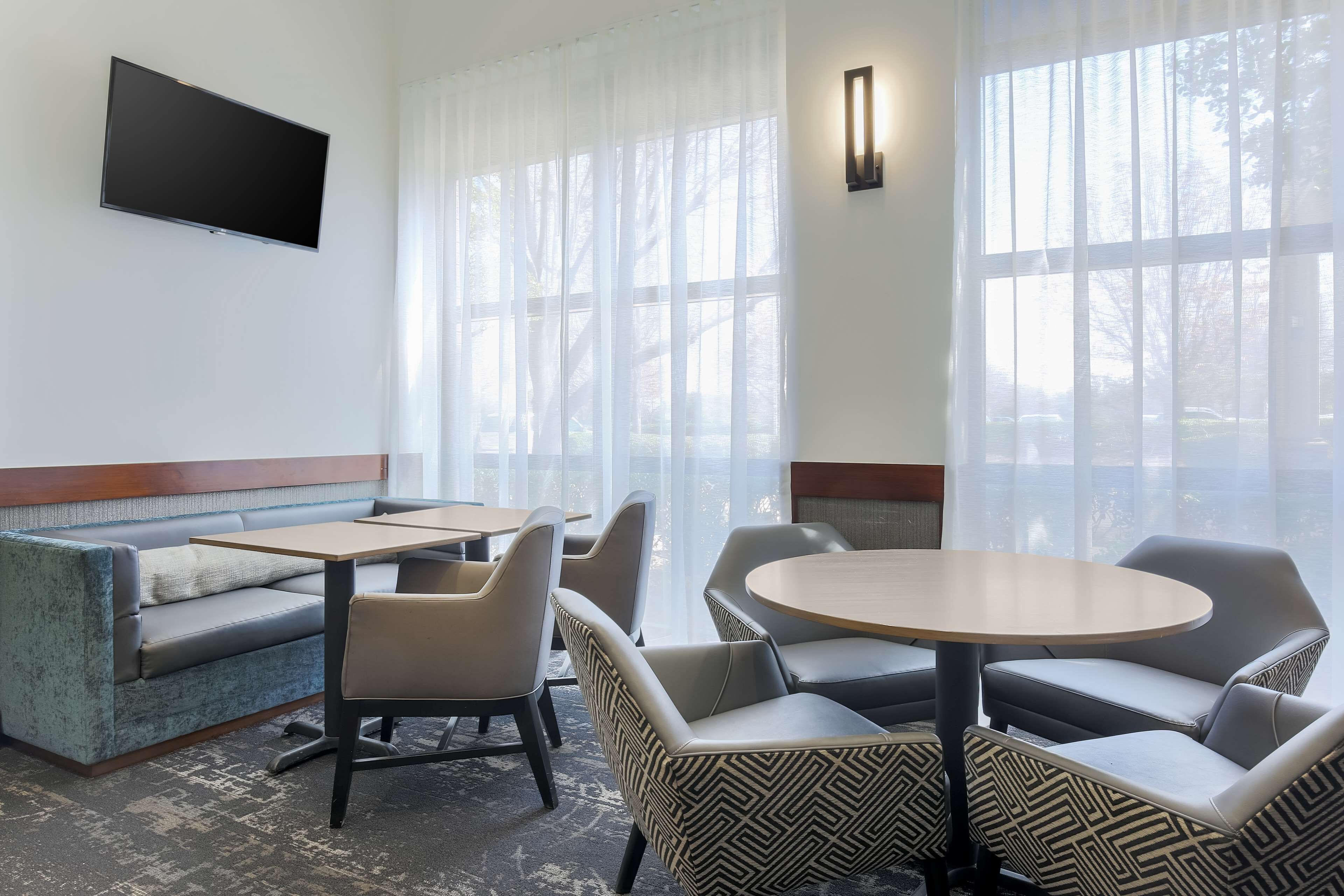 Ein Hotelzimmer mit einer Couch, Sesseln, einem Tisch, einem Flachbildfernseher und großen Fenstern mit transparenten Vorhängen.