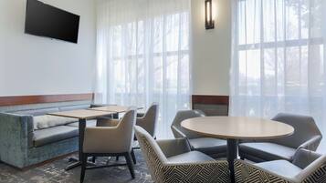 Ein Hotelzimmer mit einer Couch, Sesseln, einem Tisch, einem Flachbildfernseher und großen Fenstern mit transparenten Vorhängen.