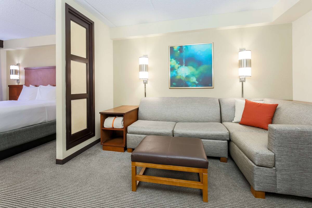A hotel room with a bed, a gray sofa, a wooden bench, and two wall-mounted lamps.