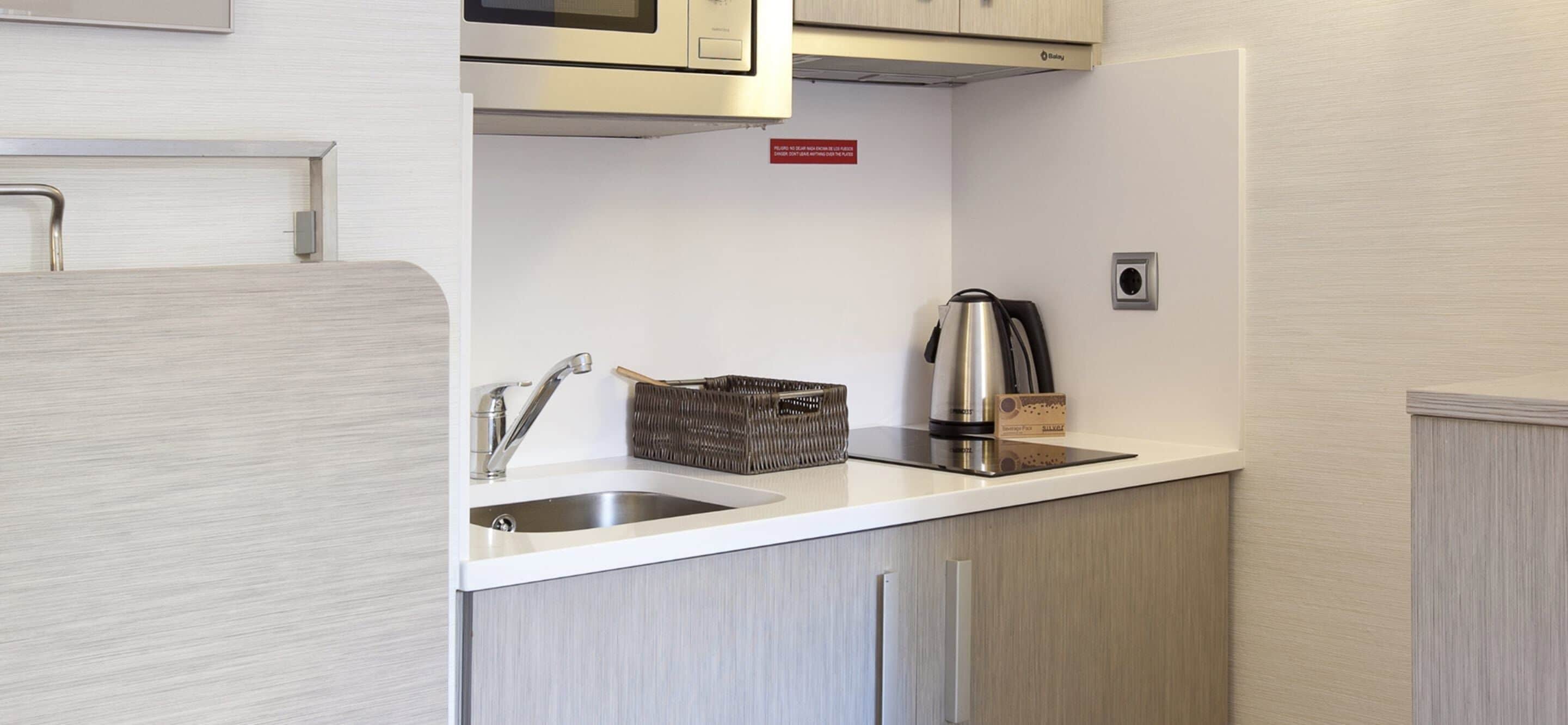 Una cocina compacta con un lavamanos de acero inoxidable, una tetera y un microondas sobre la encimera.