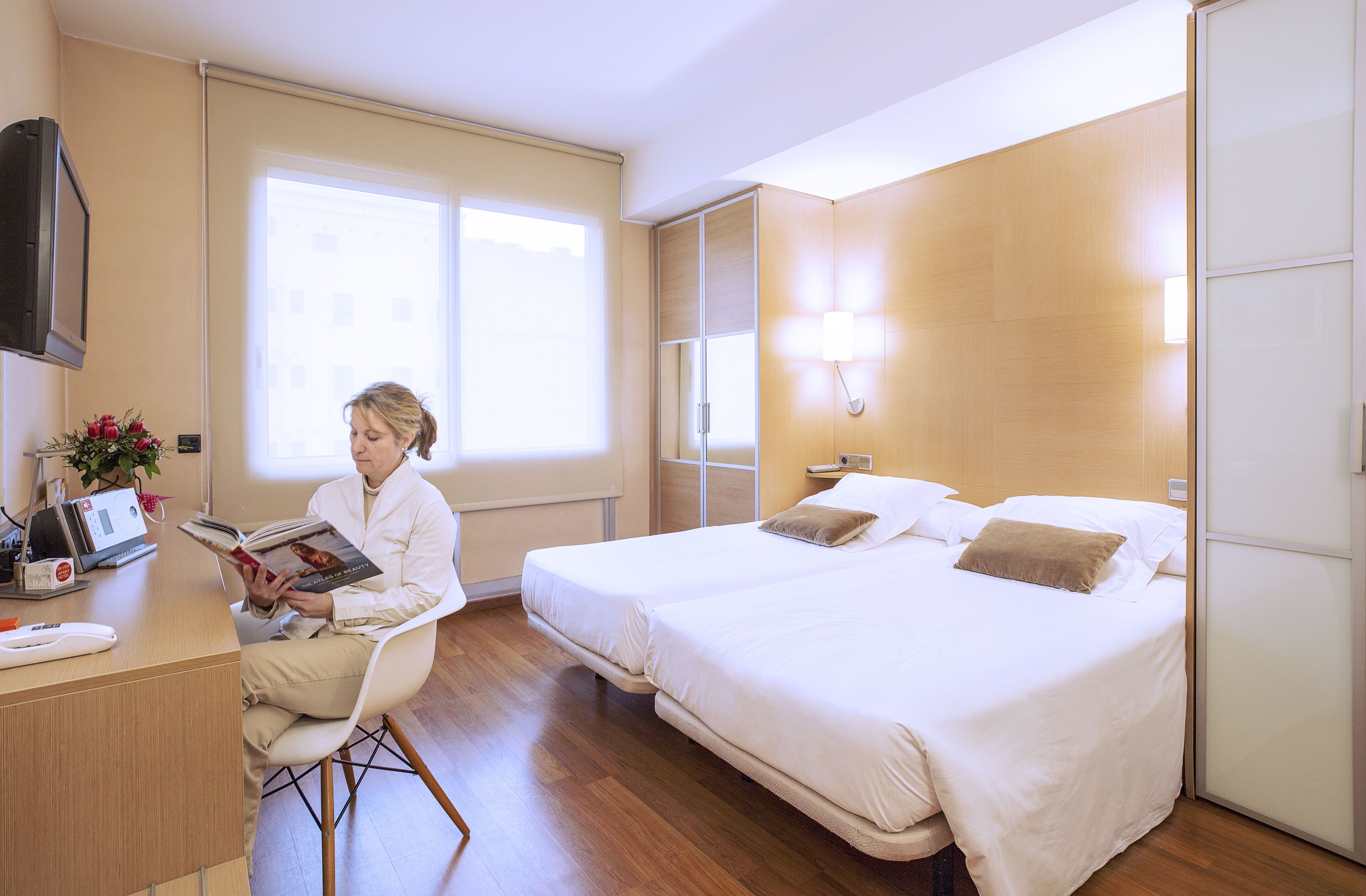 A hotel room with a bed, bedside table, lamp, and a person reading a book.