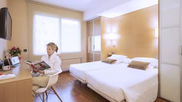 A hotel room with a bed, bedside table, lamp, and a person reading a book.