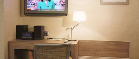 A hotel room with a television displaying a news broadcast, a desk with a lamp, a chair, and a framed picture on the wall.