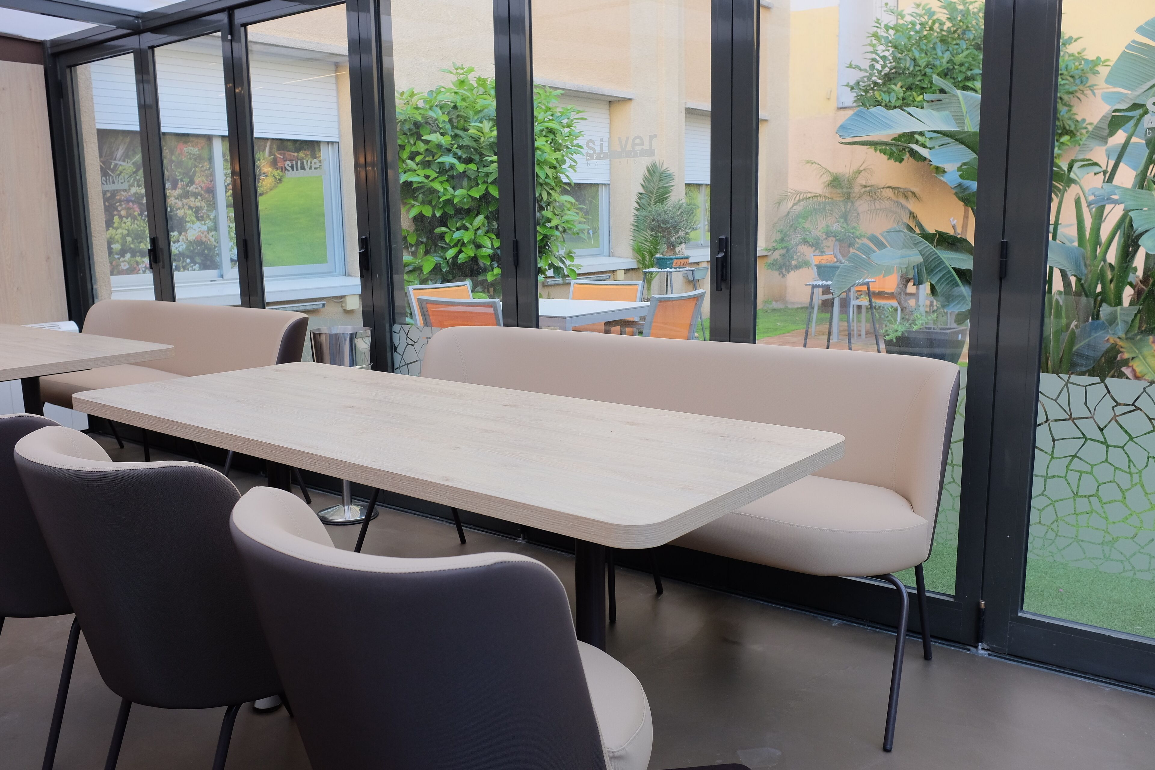 A modern indoor space with a long table and several chairs, large windows offering a view of a garden and neighboring buildings.