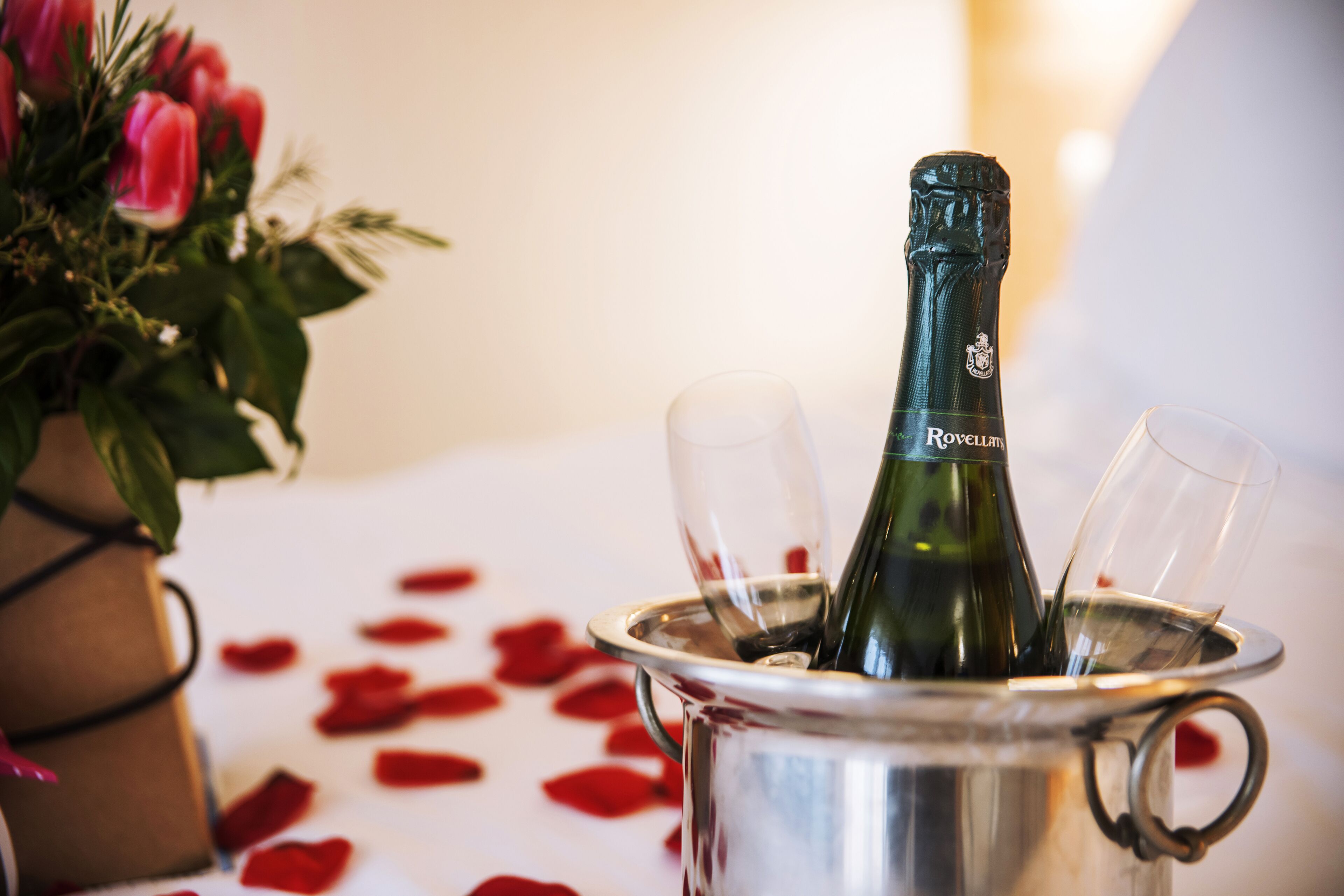 Una bottiglia di champagne in un secchio di metallo con due bicchieri nelle vicinanze, circondato da petali di rosa.