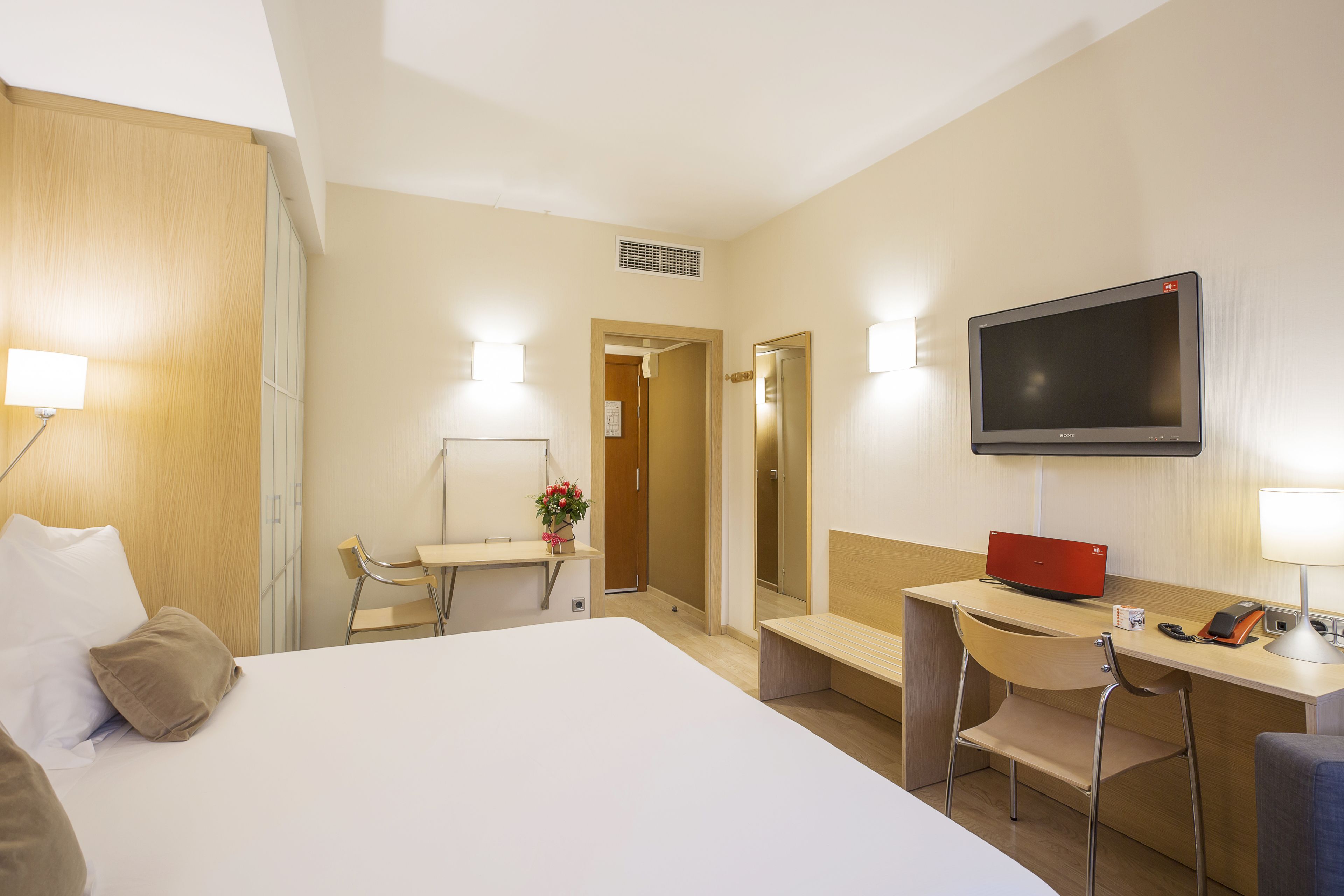 A hotel room with a bed, desk, chair, TV, and a small table with a lamp.