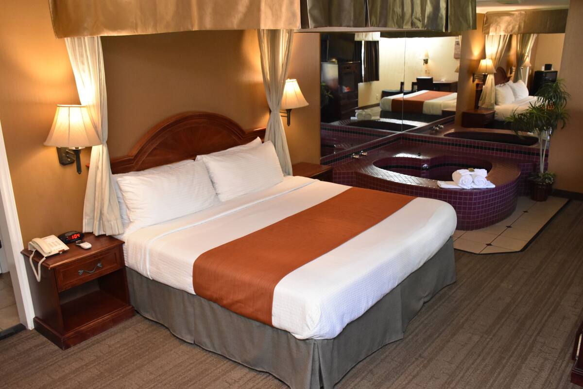 Habitación de hotel con una cama grande, una mesita de noche de madera con un teléfono, un baño con bañera y un espejo que refleja la habitación.