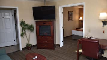 A hotel room with a flat-screen TV mounted above a wooden cabinet, a desk with a chair, a potted plant, and a bed in the background.