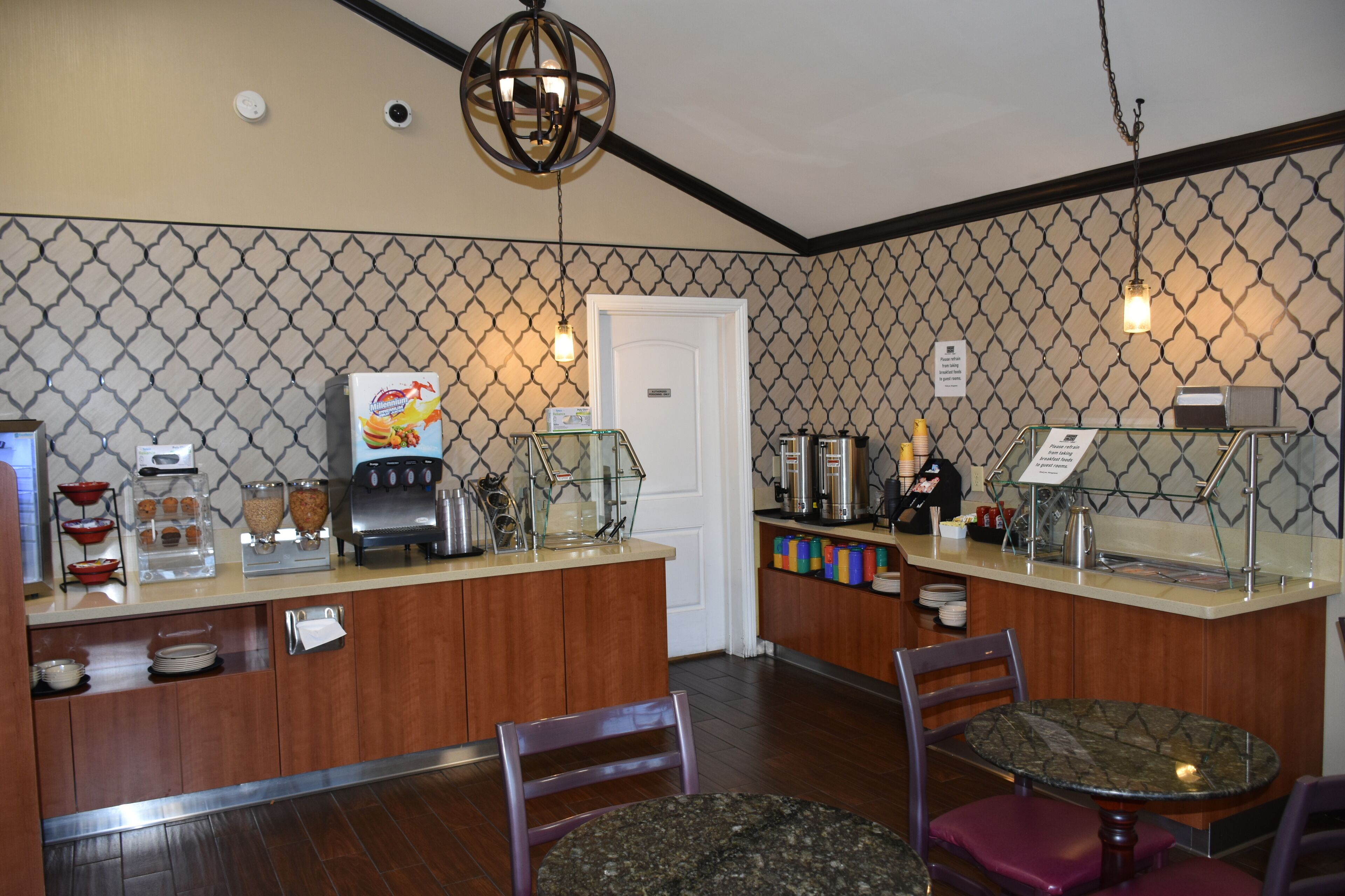 Espaço de café da manhã de hotel com máquina de café, dispensador de leite e exposição de frutas. Há duas áreas de estar com cadeiras e mesa.