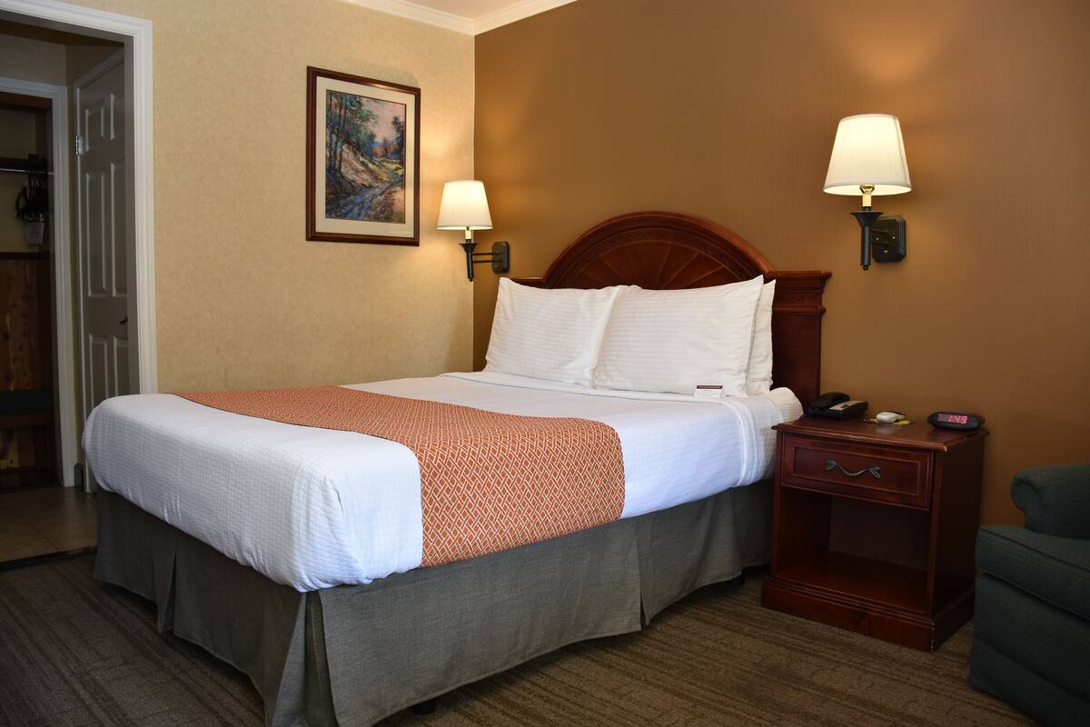 Una habitación de hotel con una cama bien hecha, dos lámparas de noche, una mesita de noche con teléfono y reloj, y un cuadro en la pared.
