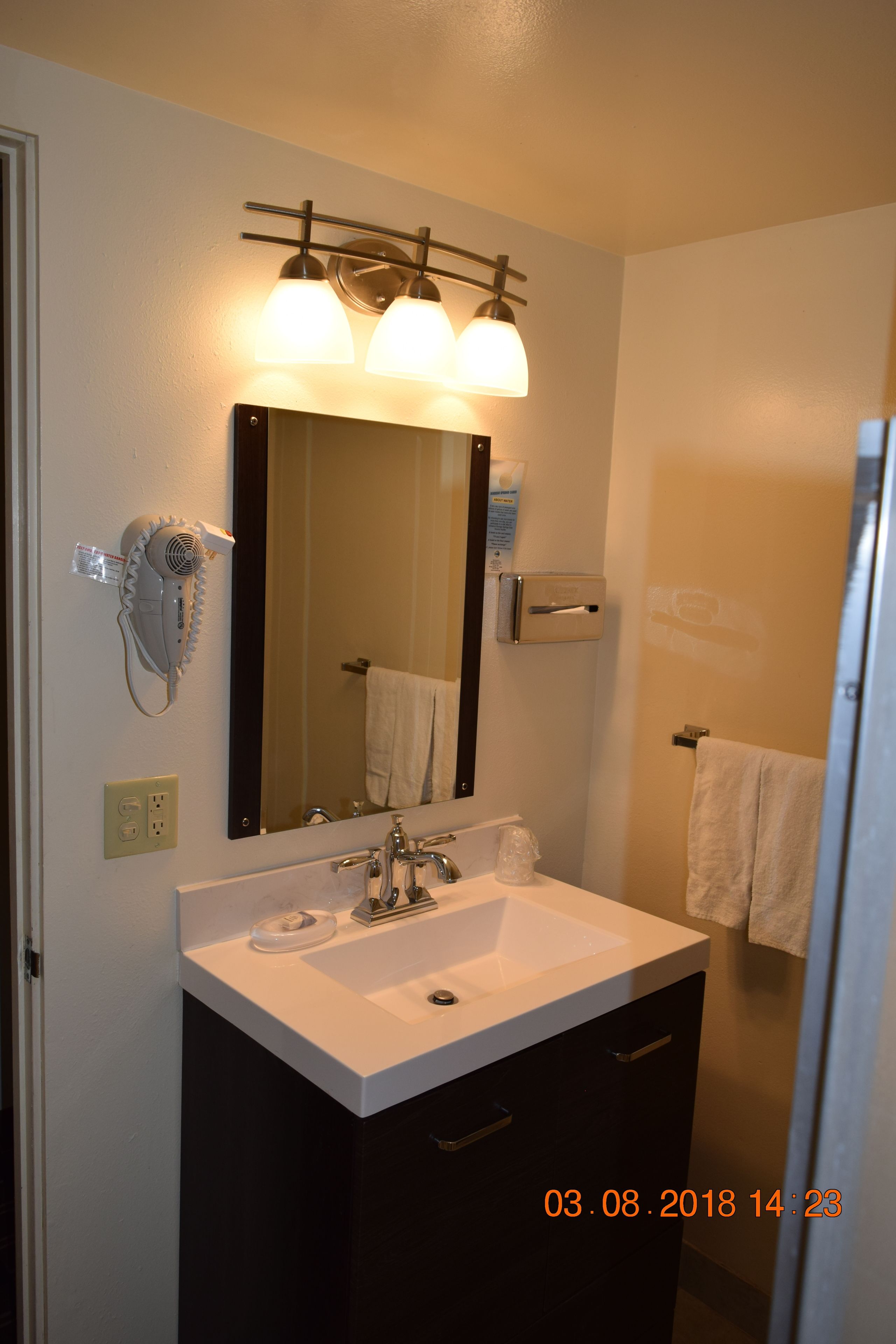 Une salle de bain avec un lavabo blanc, des meubles sombres, un miroir et un sèche-cheveux fixé au mur.