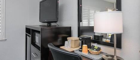 A hotel room with a desk, a television, a mirror, a lamp, and a small kitchenette area.