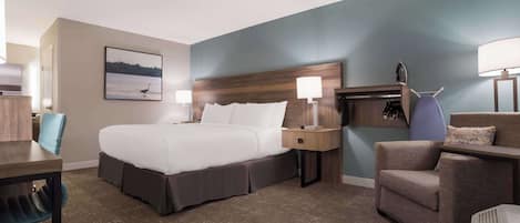 Habitación de hotel con una cama grande, dos mesitas de noche con lámparas, un escritorio con una silla y un sillón con un cojín.