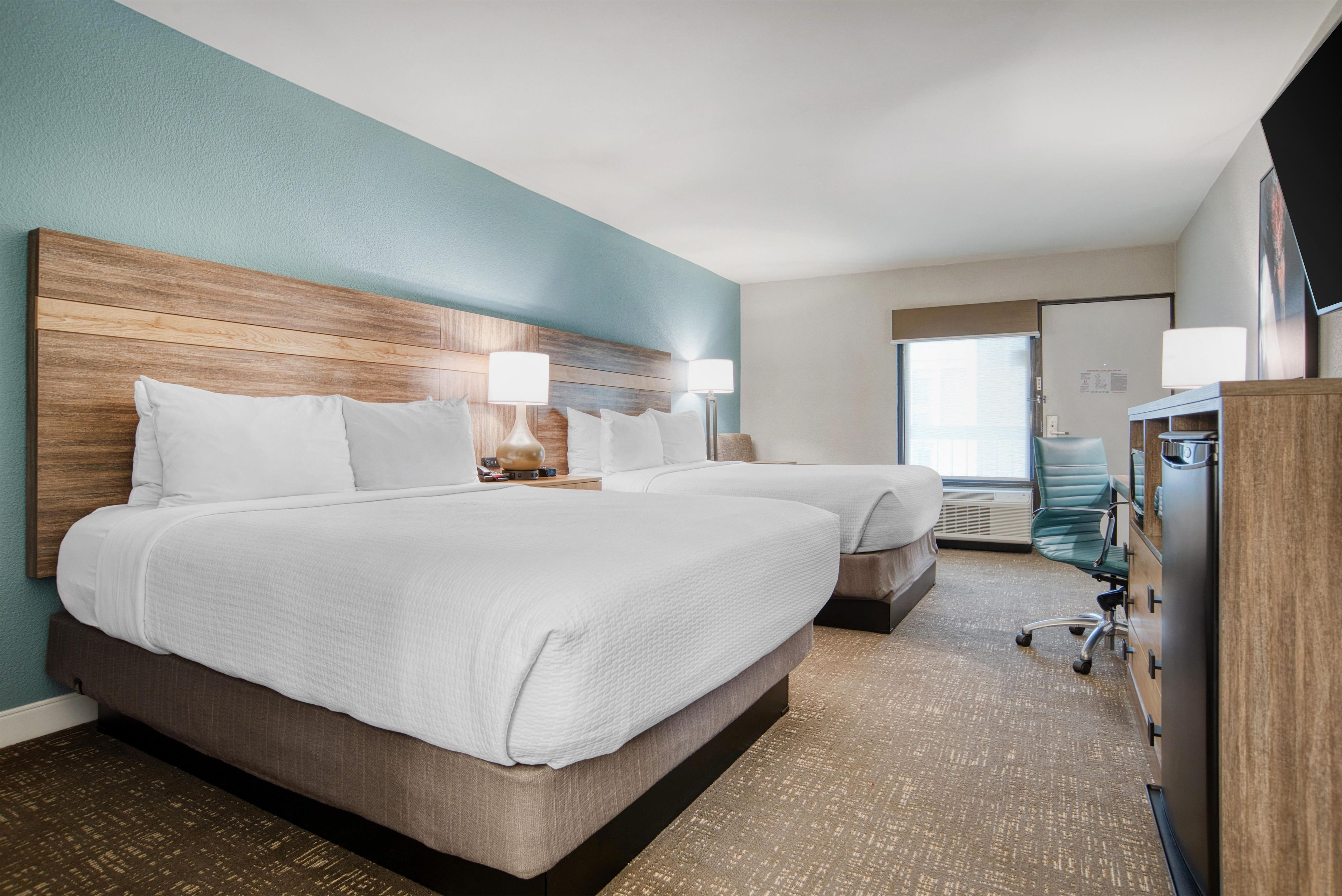 A hotel room with two beds, a wooden headboard, a desk with a lamp, and a view of the city through the window.