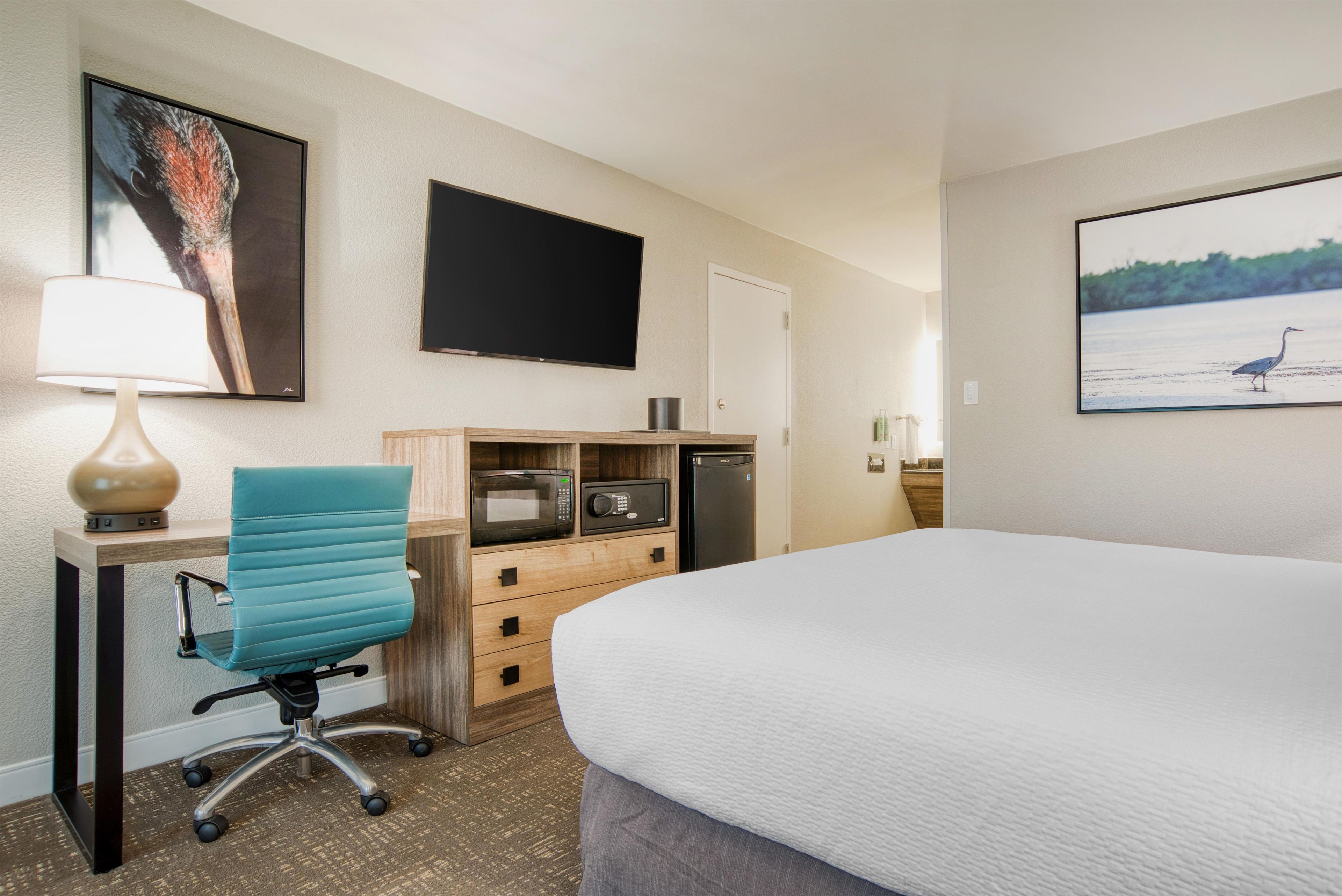 A hotel room with a bed, a desk with a lamp, a chair, a flat-screen TV, a microwave, a refrigerator, and a painting on the wall.