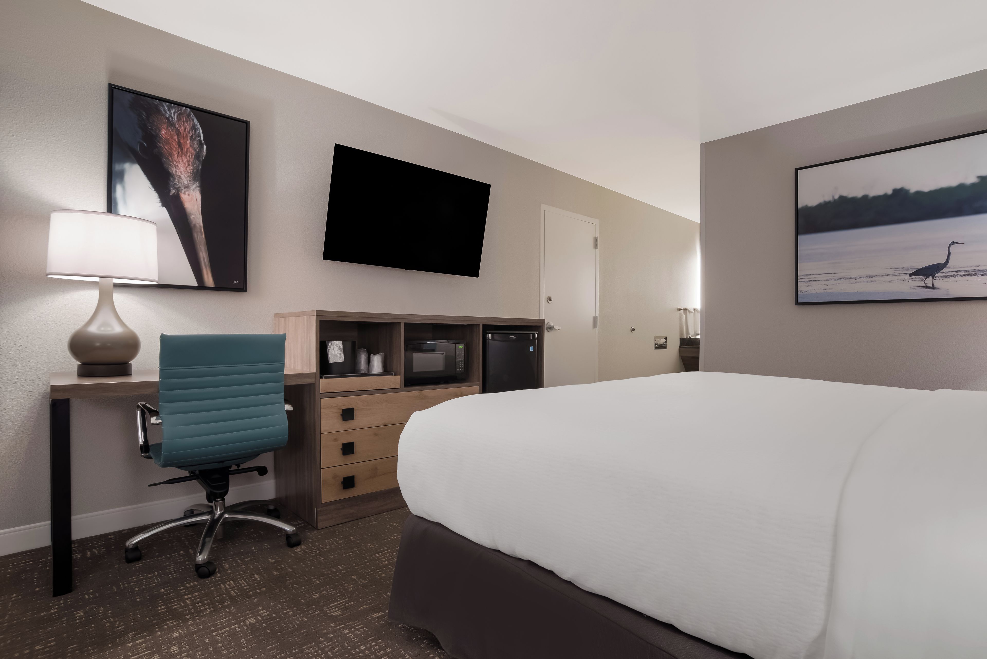 A hotel room with a bed, desk, chair, flat-screen TV, and artwork on the walls.