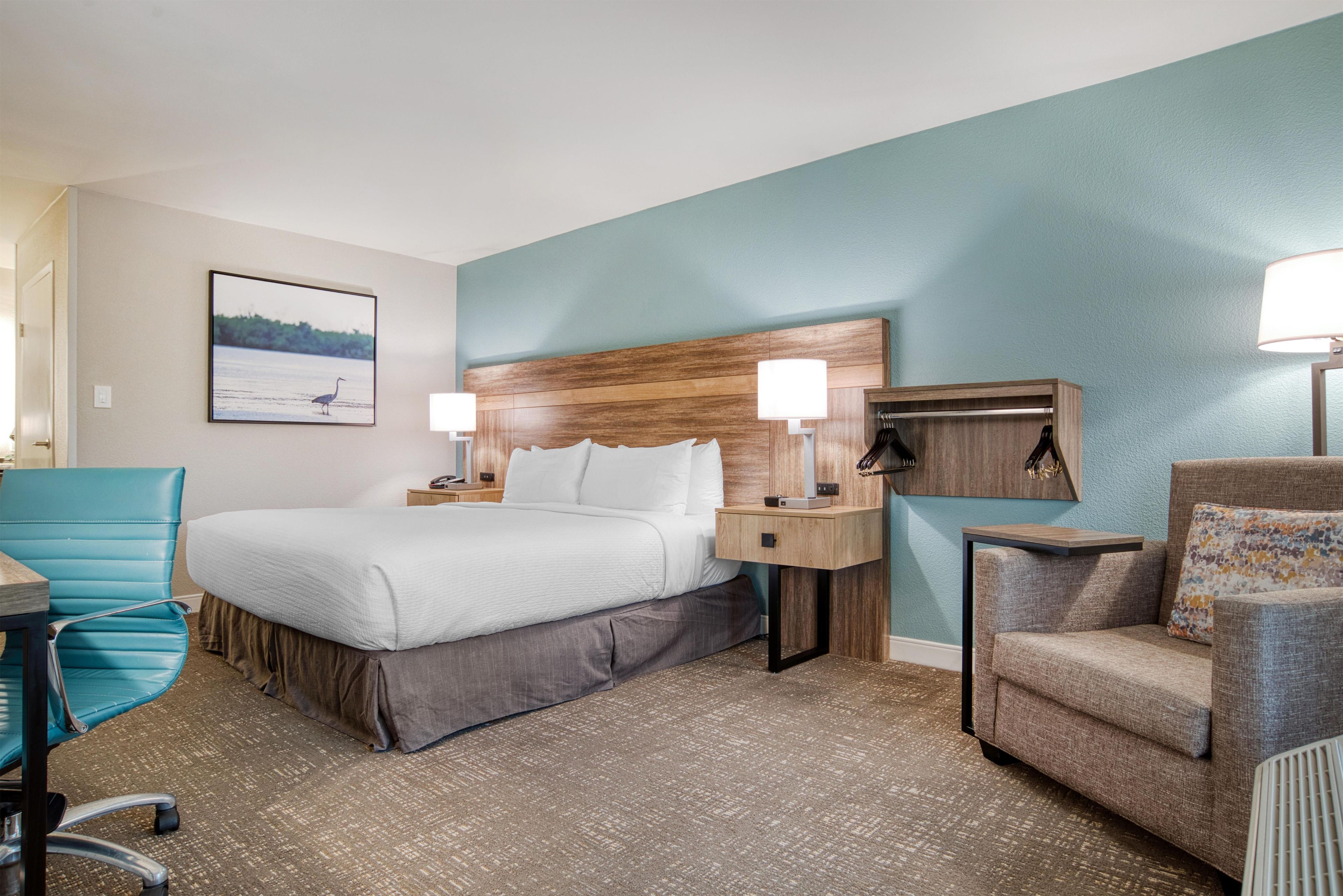 A hotel room with a bed, a desk, a chair, a lamp, and a wall-mounted shelf.