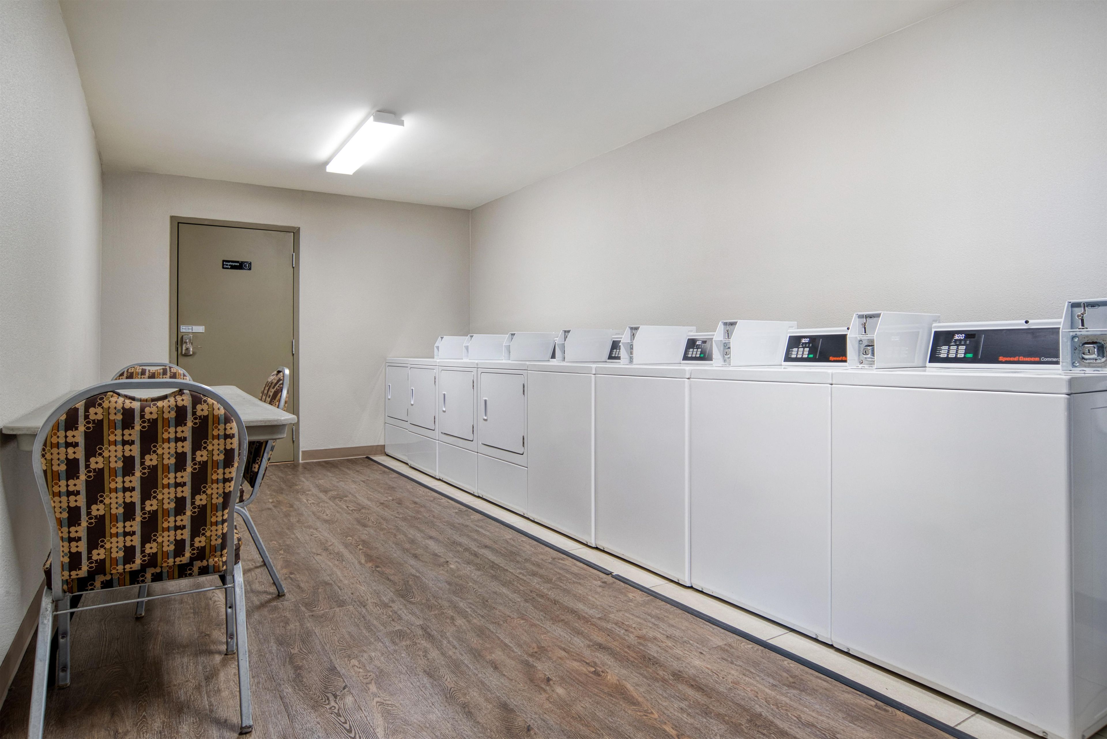 Une buanderie avec une rangée de machines à laver blanches, une chaise avec un coussin à motifs et une porte portant l’inscription « Réservé au personnel ».