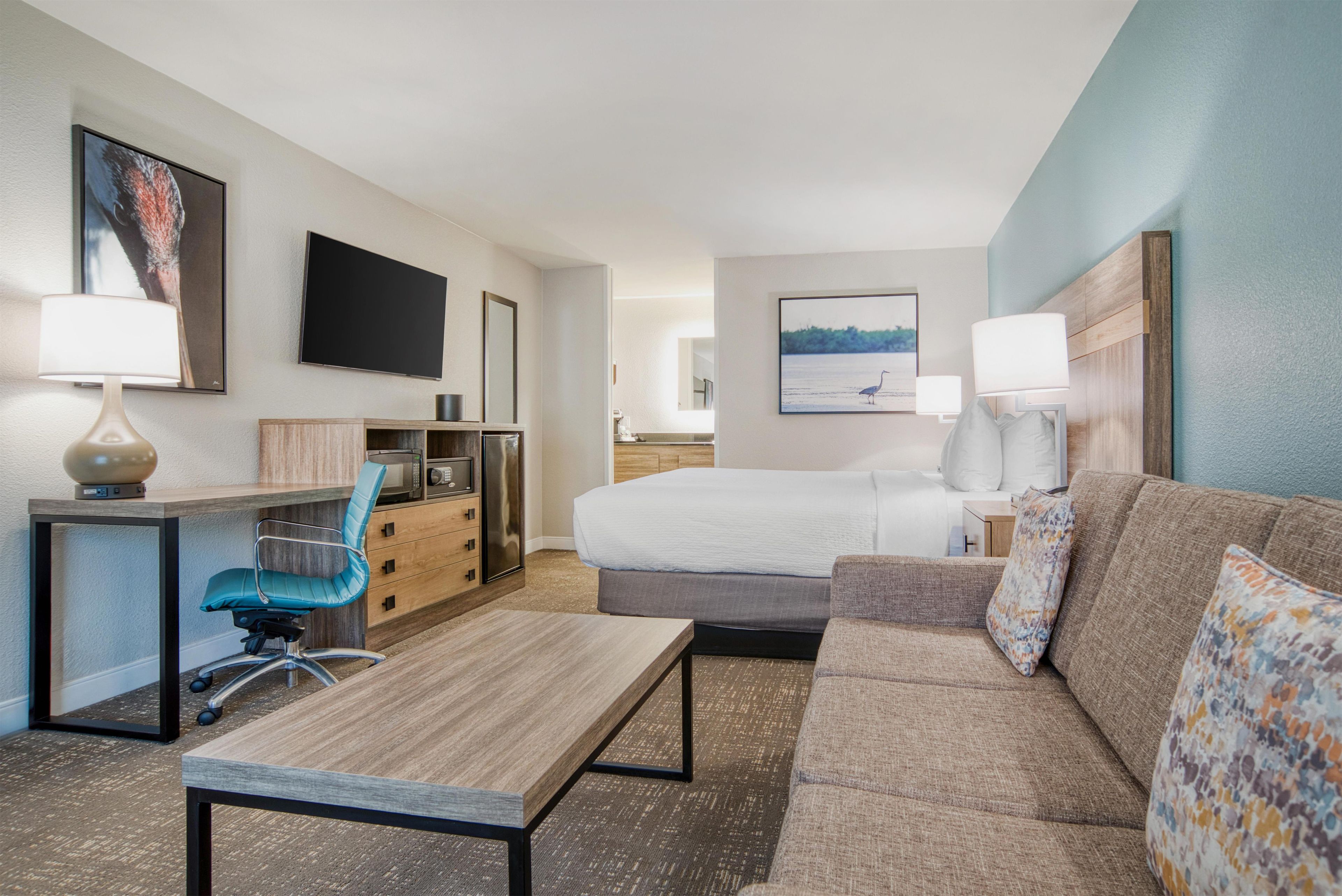 A hotel room with a bed, a desk with a chair, a sofa, a TV, and artwork on the walls.