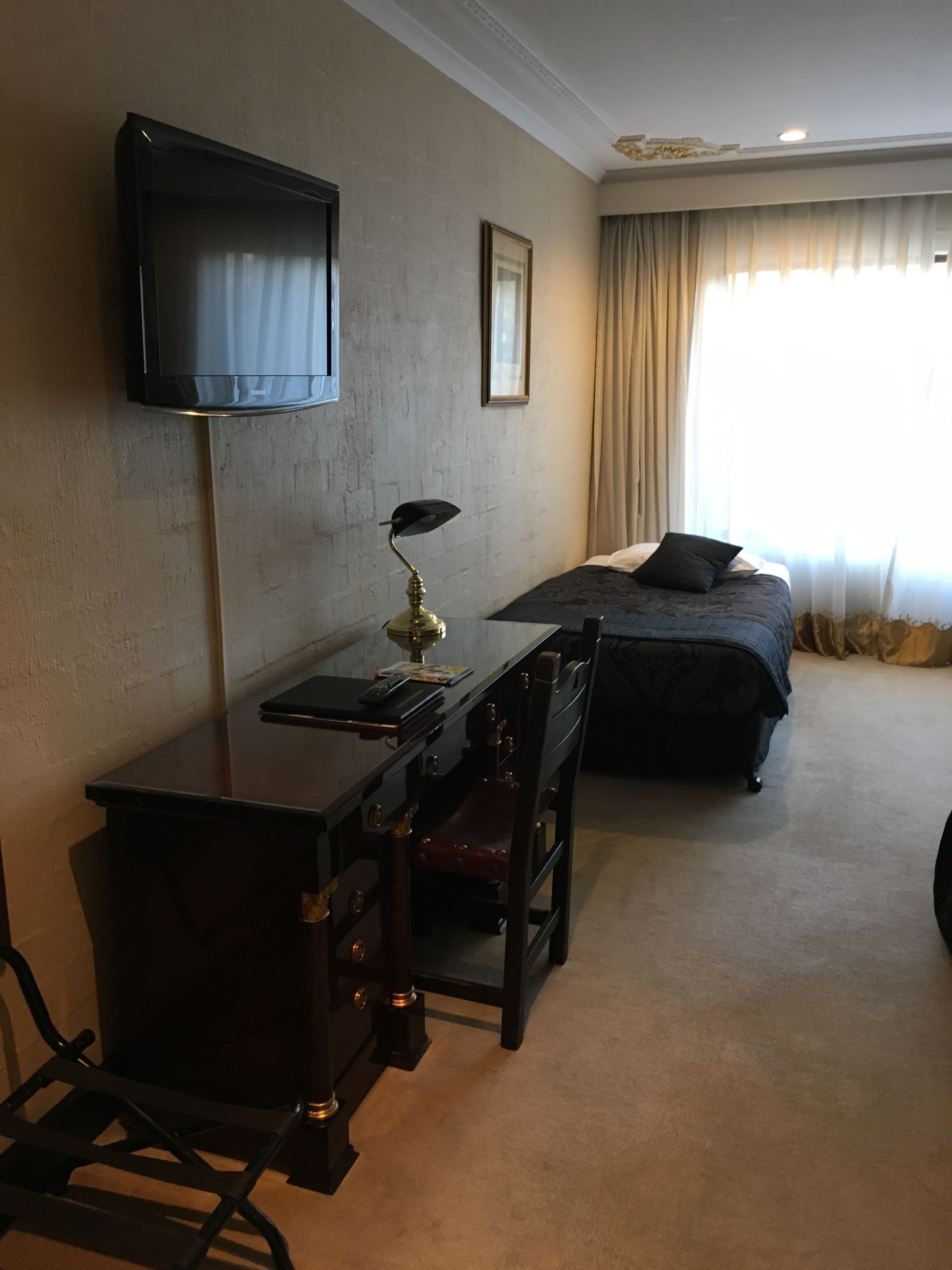 A hotel room with a bed, a desk with a chair, a television mounted on the wall, and a window with curtains.
