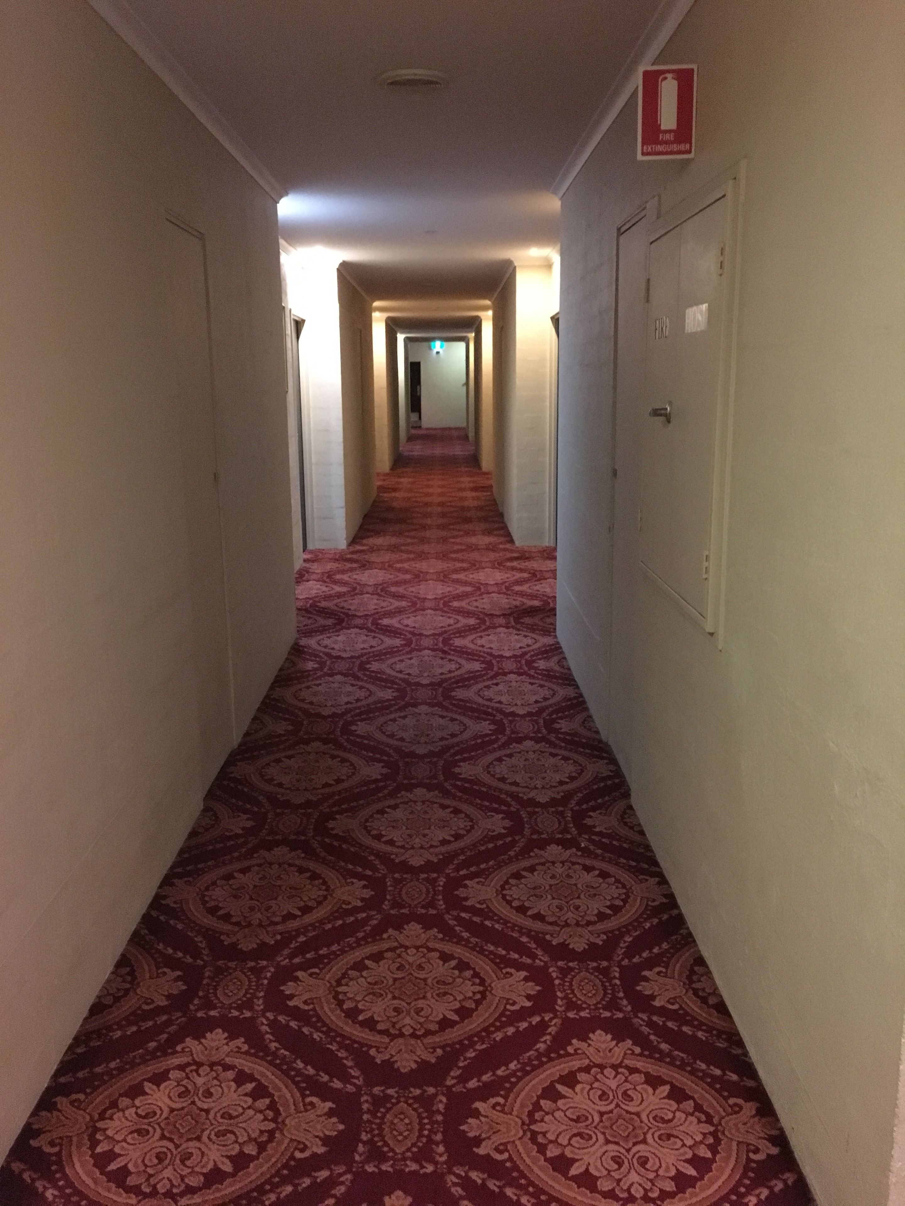 A hotel hallway with patterned carpet, doors on both sides, and a fire extinguisher sign on the right wall.