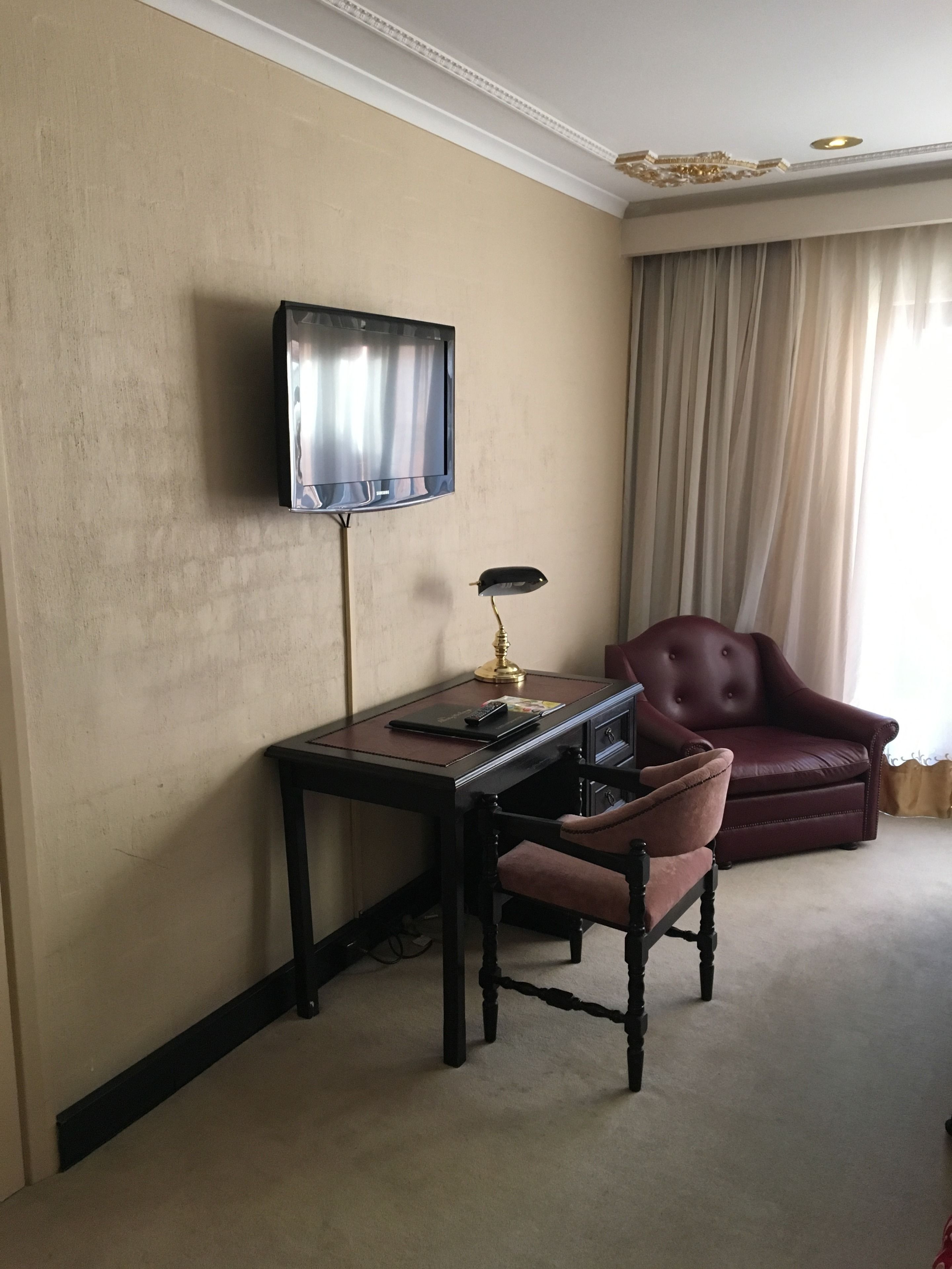 Quarto com televisão na parede, escrivaninha com abajur, poltrona de couro e uma janela grande com cortinas.