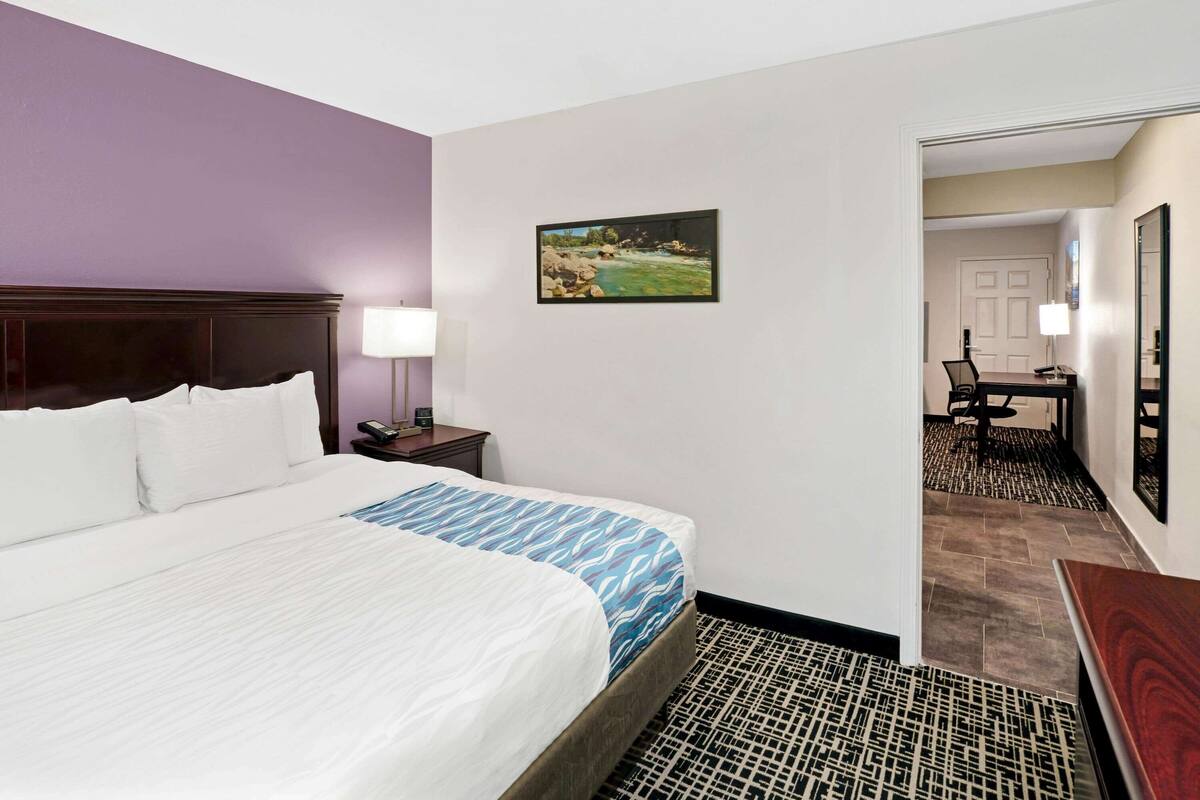 A hotel room with a large bed, a nightstand, a lamp, a painting, and a desk with a chair.