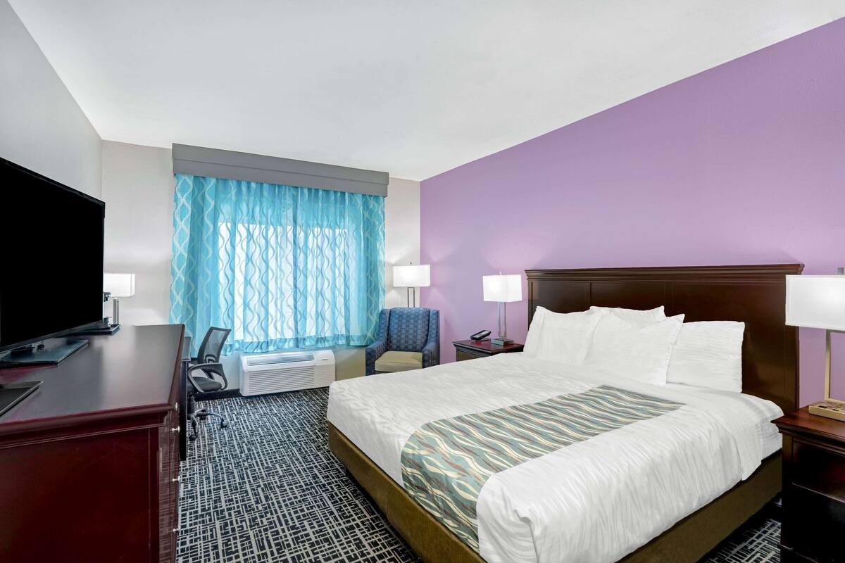 A hotel room with a large bed, a desk with a TV, a chair, a desk lamp, and a window with curtains.