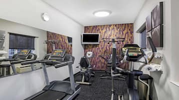 A gym with treadmills, elliptical machines, and a stationary bike. There is a TV mounted on the wall and a window with blinds.