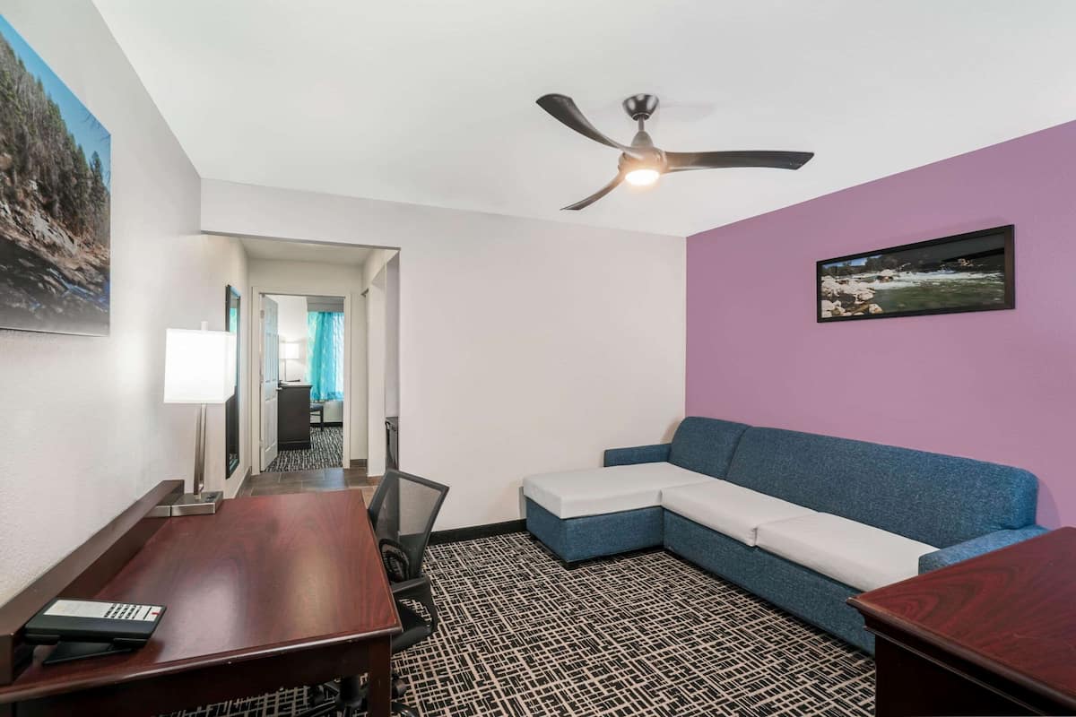 A hotel room with a ceiling fan, a sofa, a desk with a keyboard, and a patterned carpet.
