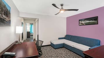 Quarto de hotel com ventoinha no teto, sofá, secretária com teclado e carpete estampado.