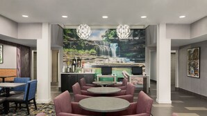A modern hotel lobby with round tables and comfortable seating, a waterfall mural on the wall, and a reception desk with a TV. - La Quinta Inn & Suites by Wyndham Cookeville (Cookeville)