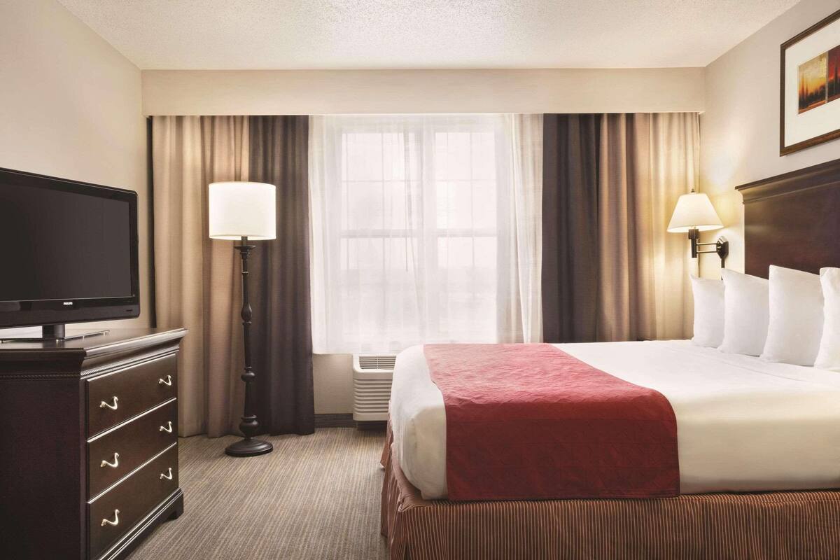 A hotel room with a bed, a TV, a dresser, a lamp, and a window with curtains.