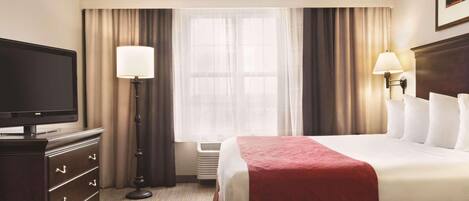 Habitación de hotel con cama, televisor, cómoda, lámpara y ventana con cortinas.