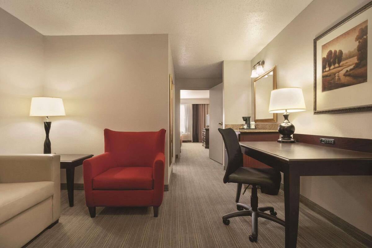 A hotel room with a red armchair, a desk, a chair, a lamp, and a painting on the wall.