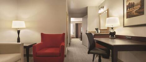Una habitación de hotel con un sillón rojo, un escritorio, una silla, una lámpara y un cuadro en la pared.