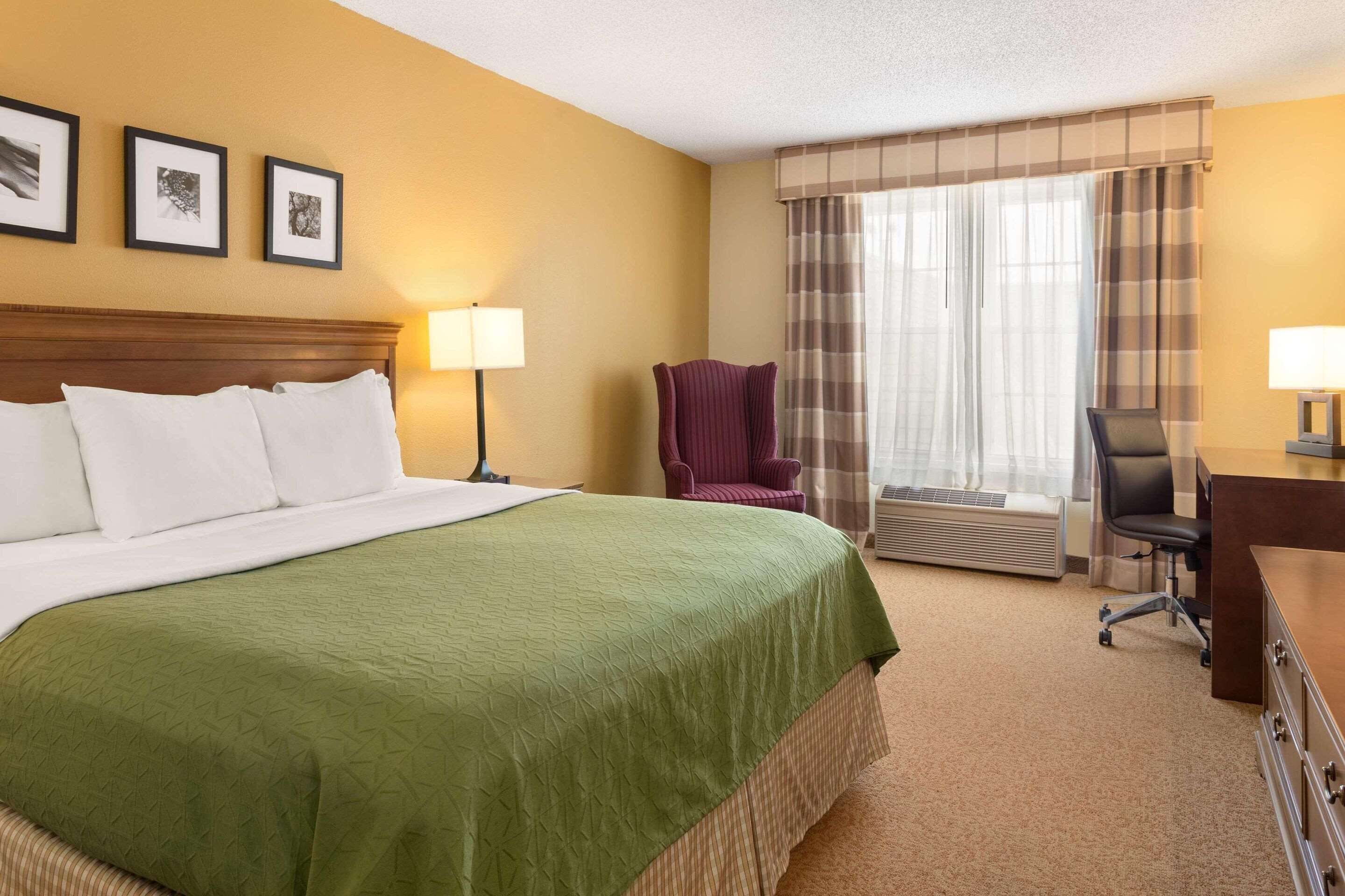Una habitación de hotel con una cama grande, un escritorio, una silla, una lámpara y un ventanal con cortinas.
