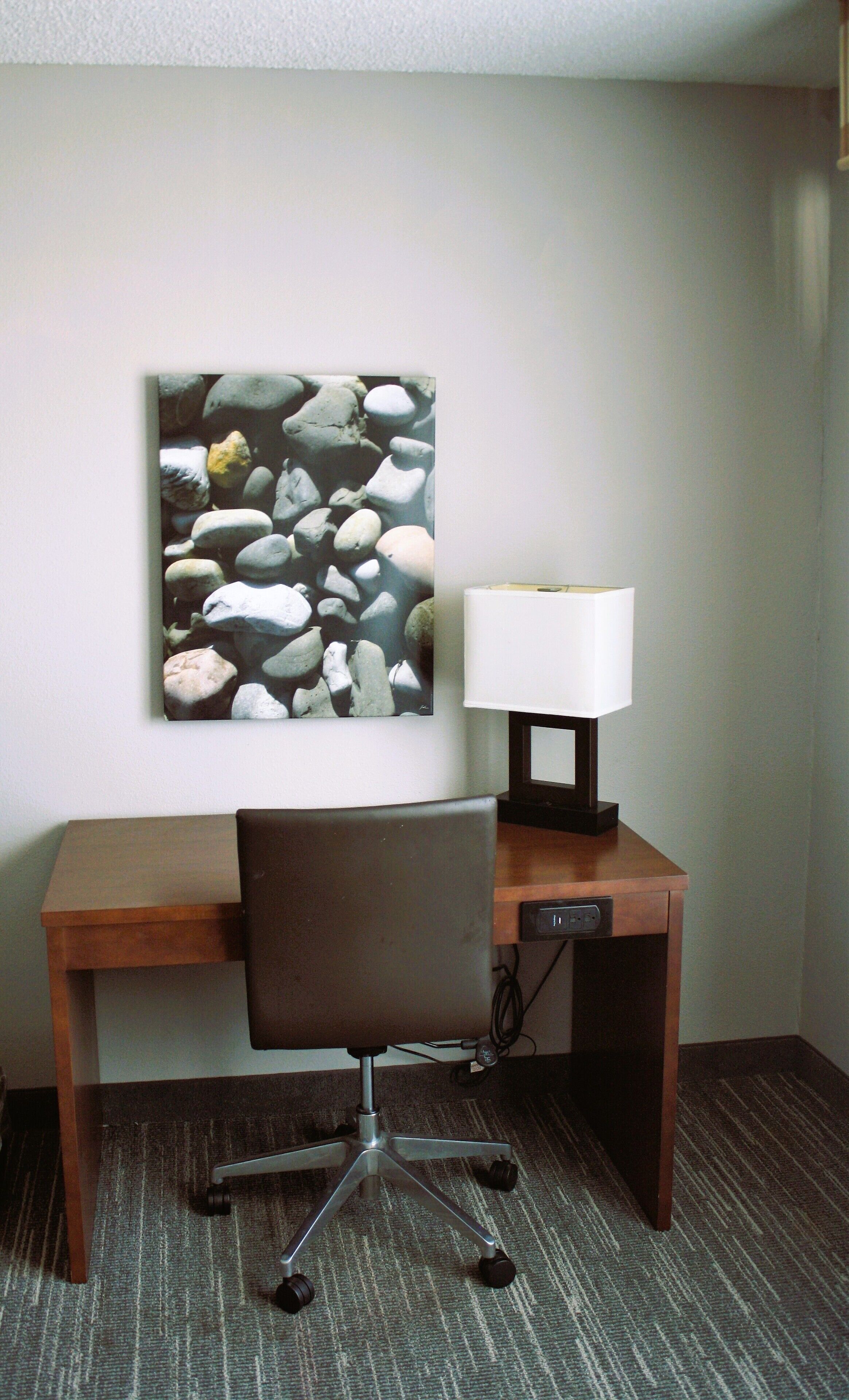 Habitación de hotel con un escritorio de madera, una silla de oficina moderna, una lámpara y una obra de arte en la pared que representa rocas.