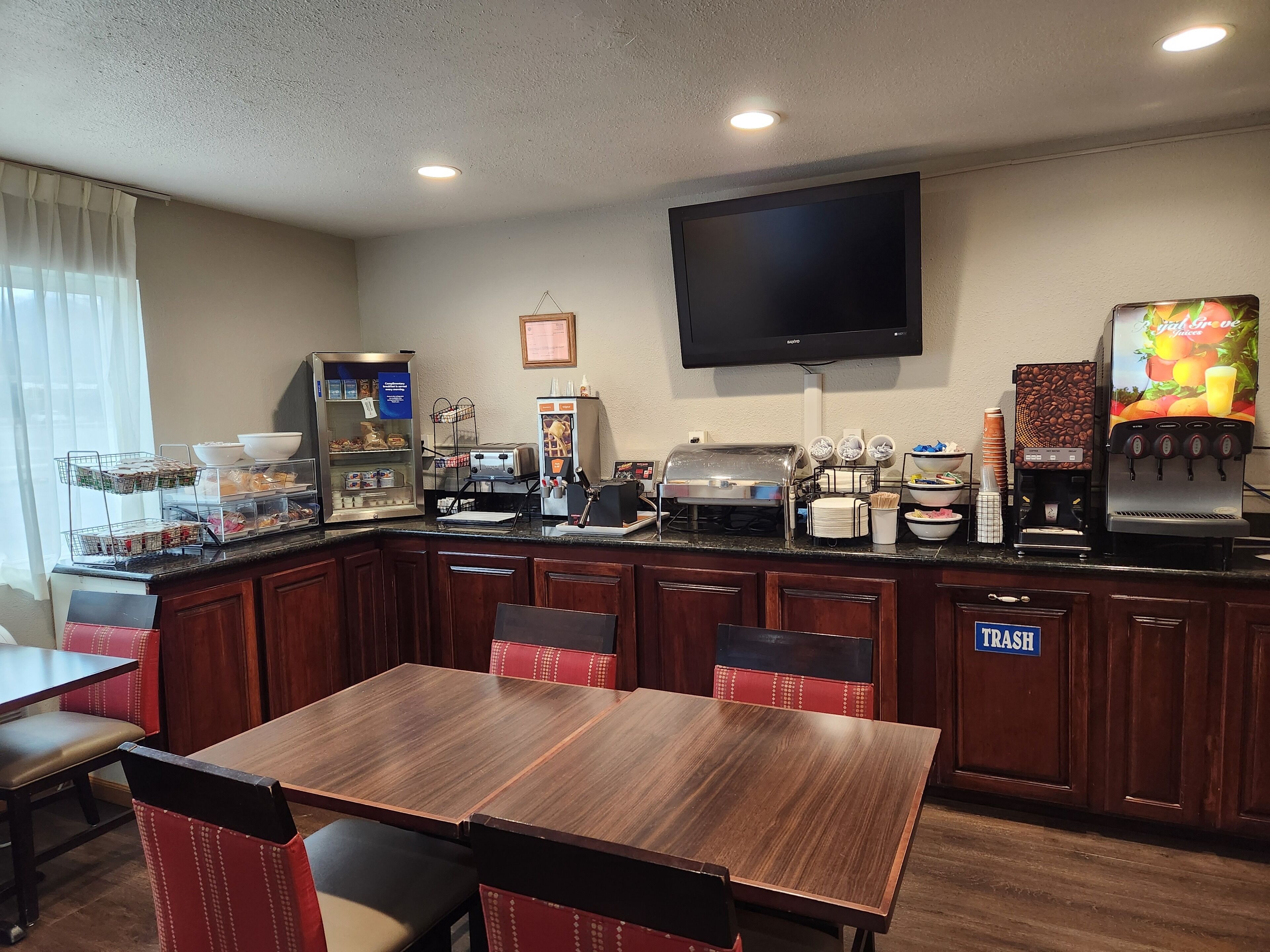 Habitació d'hotel amb una taula de dinar de fusta, cadires vermelles amb amortidor, barra de buffet amb diversos plats, una màquina de cafè i un televisor penjat a la paret.