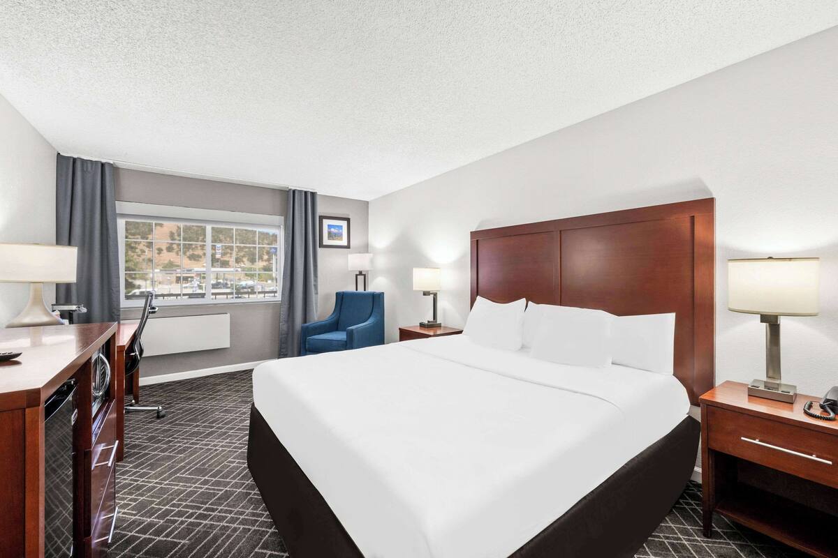 A hotel room with a large bed, two bedside tables with lamps, a desk with a chair, a blue armchair, and a window with a view of buildings.