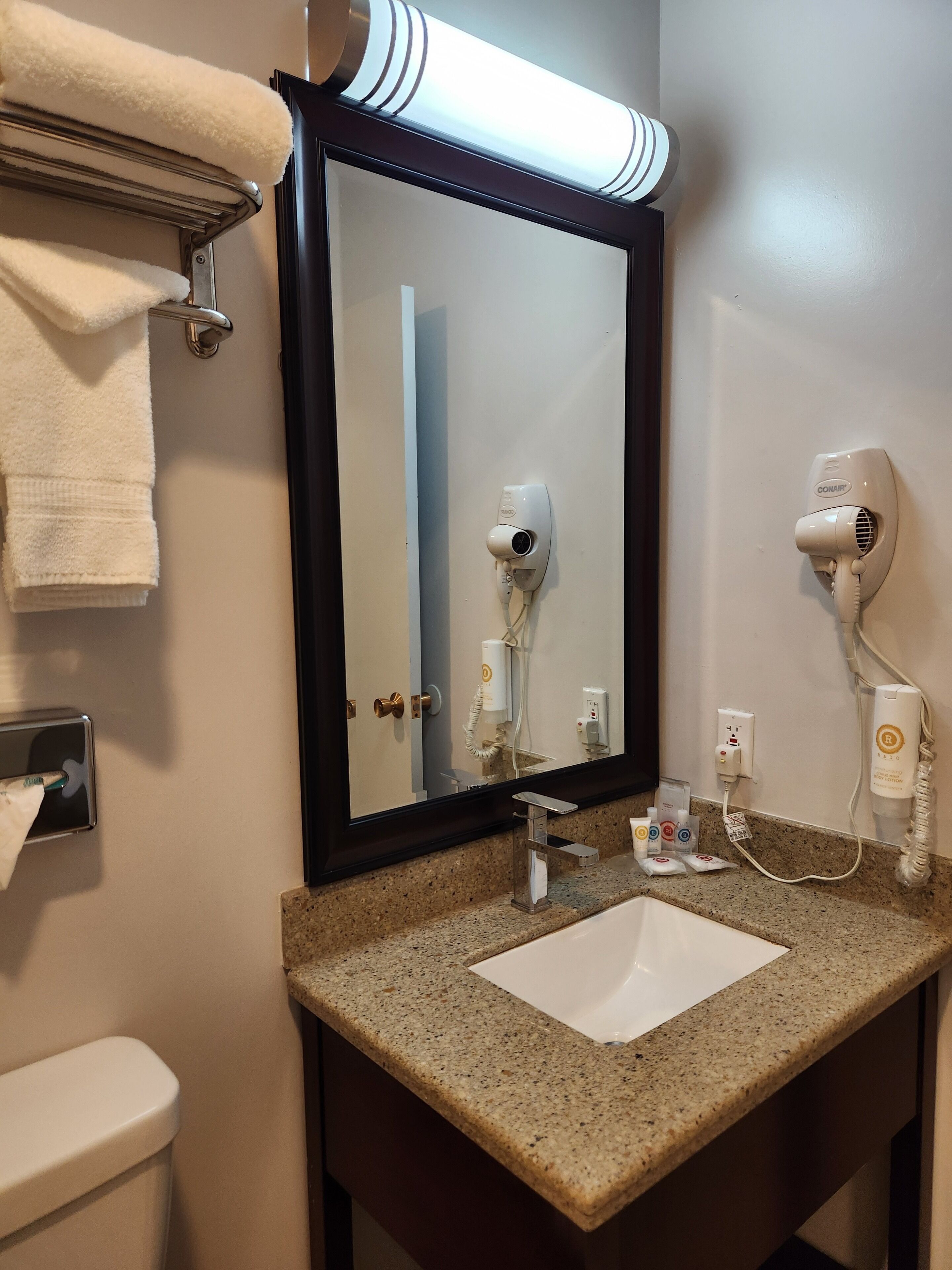 A bathroom with a granite countertop, a white sink, a mirror with a black frame, a towel rack, and two hair dryers.