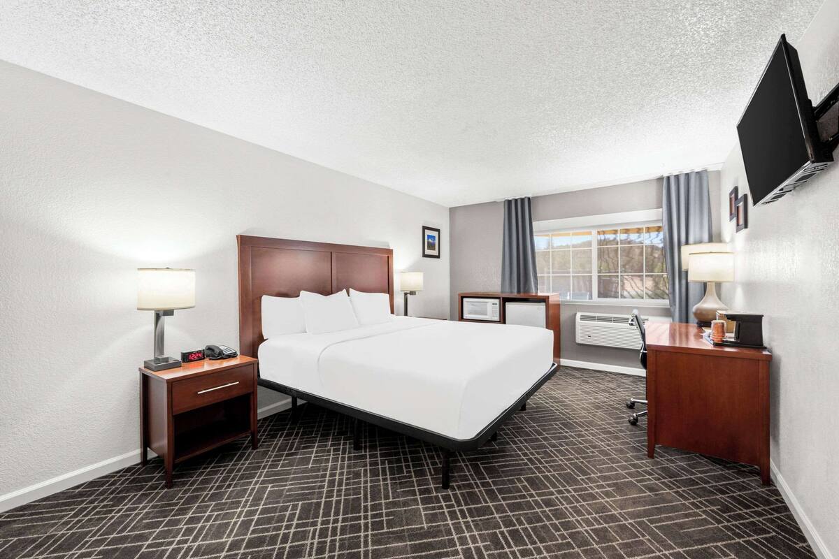 A hotel room with a bed, bedside tables, a desk, a flat-screen TV, and a window with curtains.