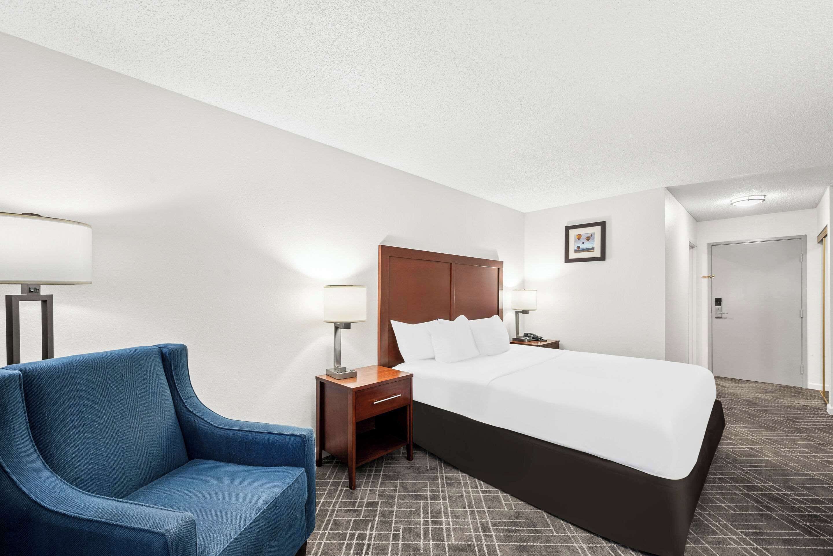 Una habitación de hotel con una cama, un sillón azul, una mesita de noche con una lámpara y un cuadro en la pared.