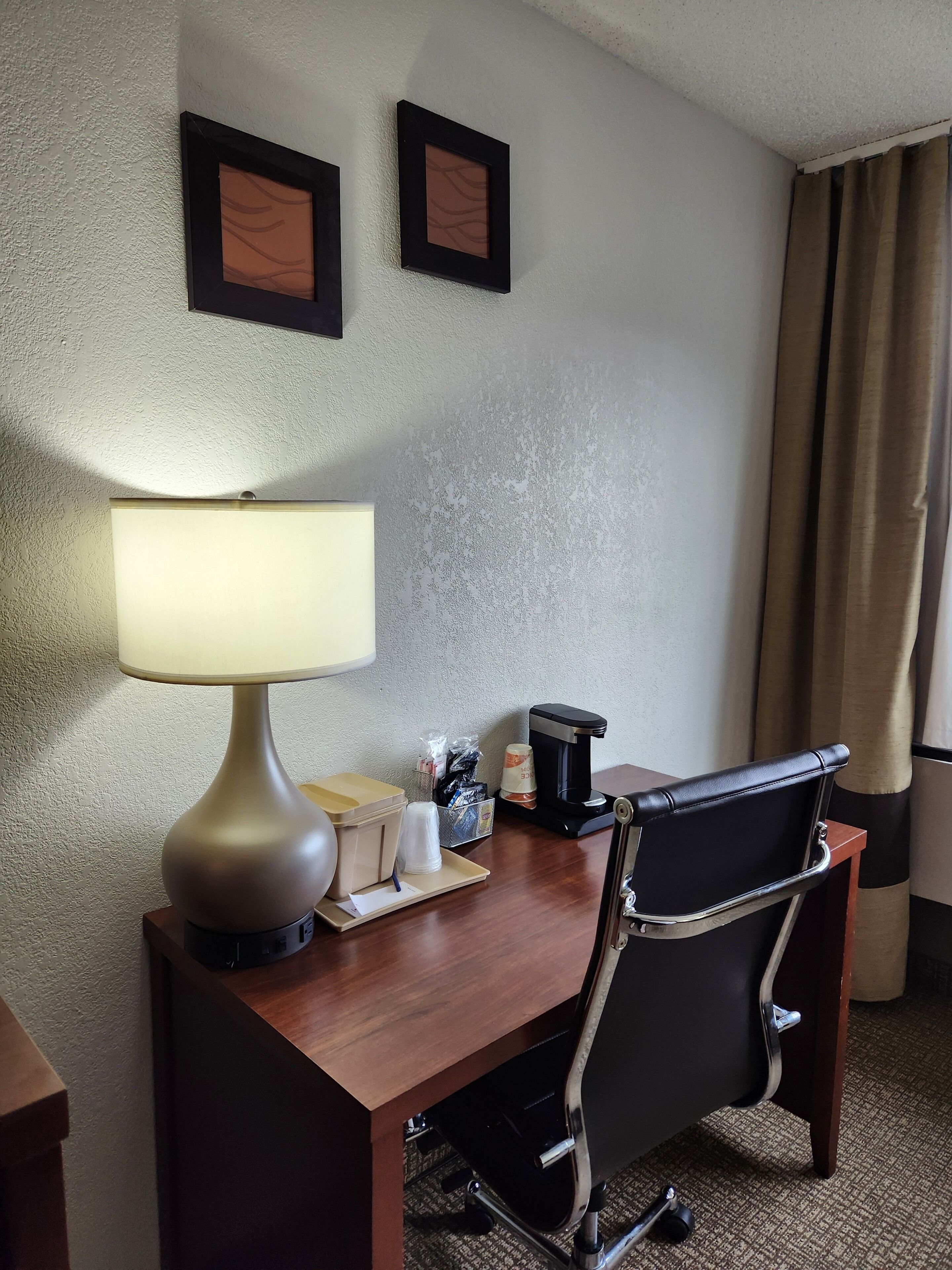 A hotel room with a desk, a lamp, a chair, a coffee maker, and two framed pictures on the wall.