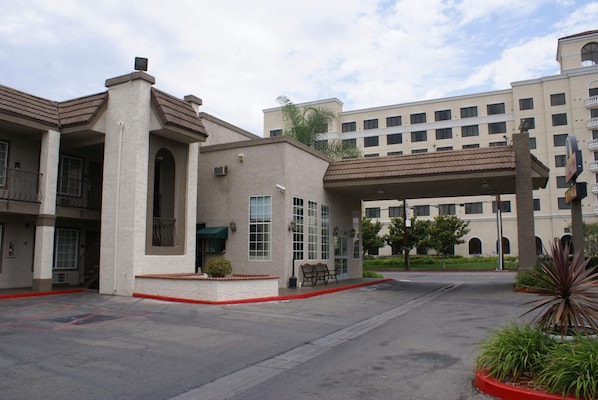 A hotel with a covered entrance, a patio area with a bench, and a parking lot. - Queens Inn Anaheim (Anaheim)