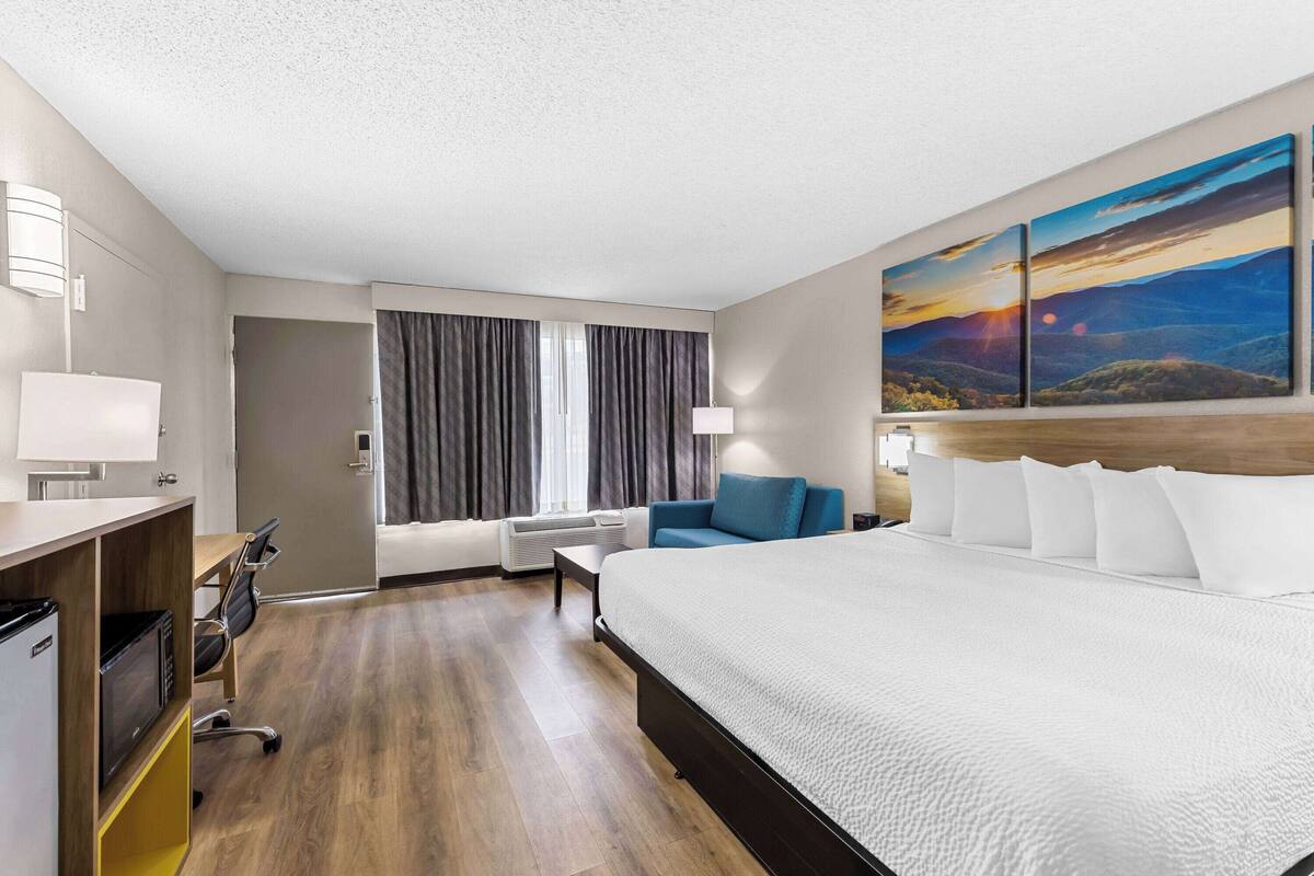 A hotel room with a large bed, a desk with a chair, a sofa, a window with curtains, and a painting on the wall.