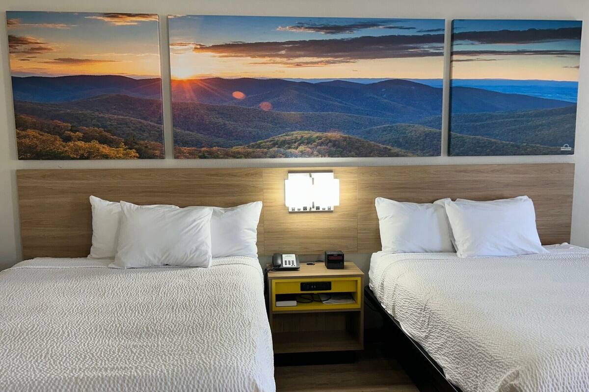 A hotel room with two beds, a wooden headboard, a nightstand with a lamp, and a wall-mounted artwork of a sunset over a mountainous landscape.